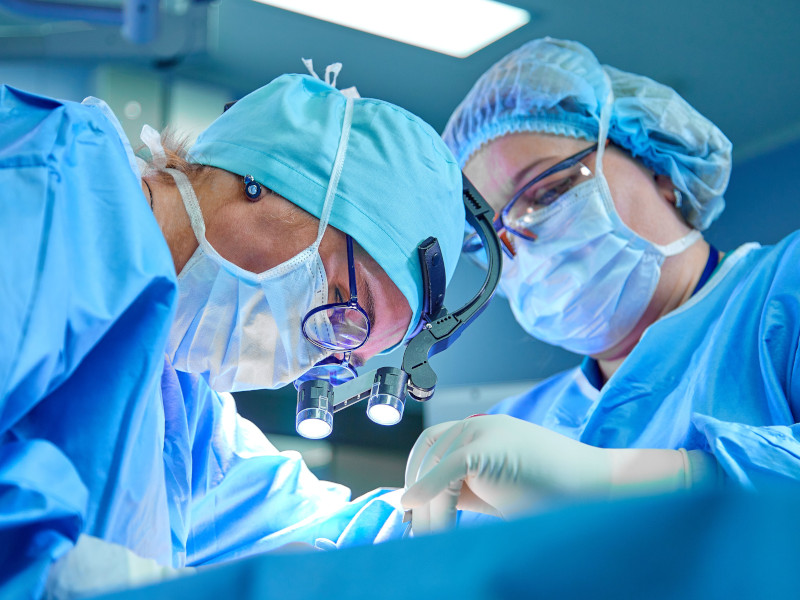 Closeup shot of two surgeons in scrubs with hair covers and facemasks looking down at patient and performing surgery