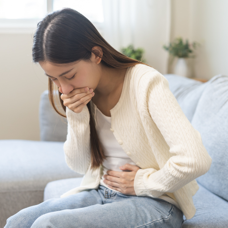 A young Asian female sitting on a couch with her hand over her mouth and other hand on her stomach looking sick