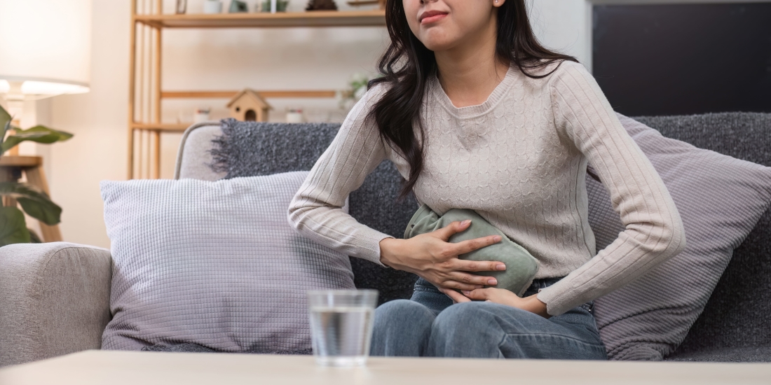 An Asian woman sitting on the coach with a heating pack on her lower abdomen, wincing from pain