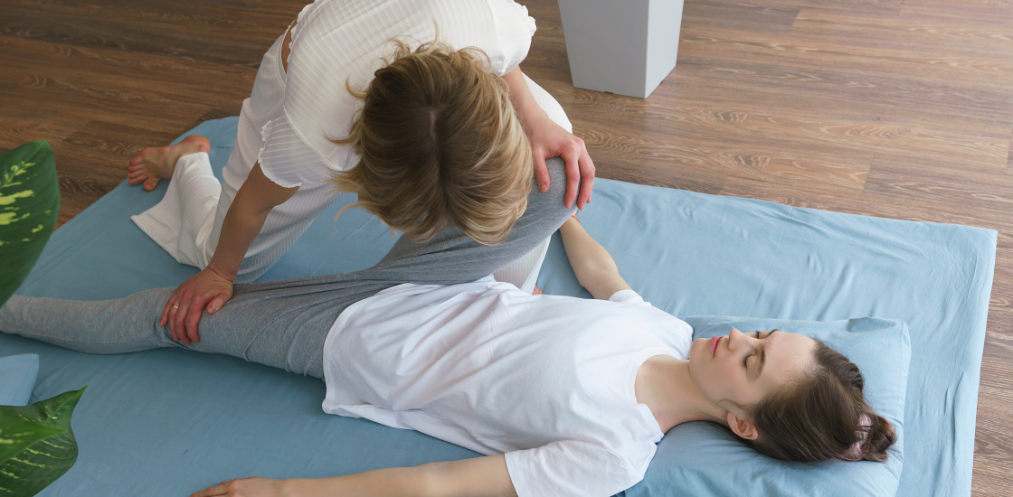 A female pelvic floor therapist performing leg and hip stretching exercises with a female patient on a yoga mat