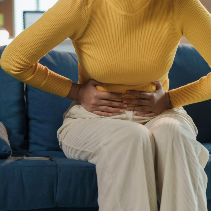 A woman sitting on a couch facing toward the camera with her hands over her lower abdomen hunched over in pain