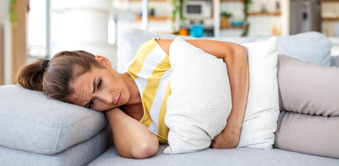 A young female laying down on a couch, in discomfort, holding a pillow tightly against her abdomen.