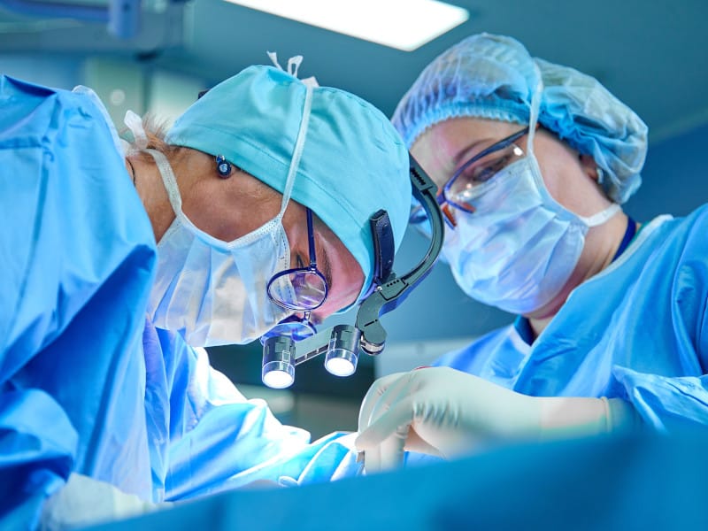 Closeup shot of two surgeons in scrubs with hair covers and facemasks looking down at patient and performing surgery