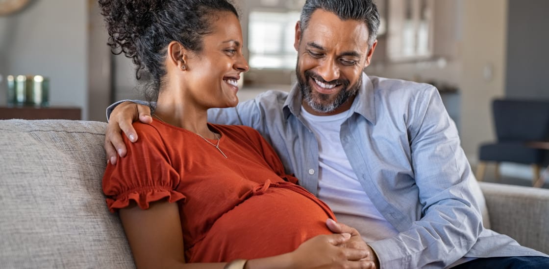 A pregnant woman sitting with her partner, both smiling with their hands on the womans belly.