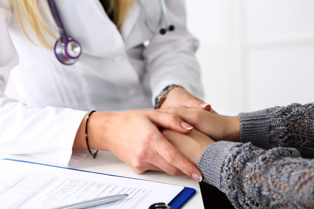 A closeup of a female doctor calmy holding the hands of a female patient in a comforting manner, a clipboard of patient intake questions on the table beside them.