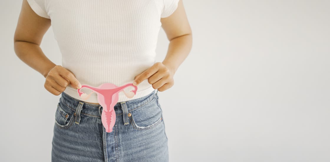 A woman standing in front of a white backdrop holding a cutout of a uterus in front of her pelvis.
