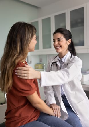 A female doctor sitting beside a female patient in an exam room with her hand on the patients shoulder offering comfort and reassurance, while patient and doctor both smile.
