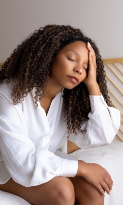 A closeup of a distressed woman sitting on the edge of her bed, hand over part of her face with eyes closed