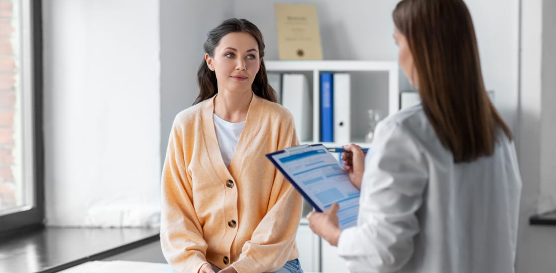 A young female sitting in a doctors office speaking with a female doctor answering questions.